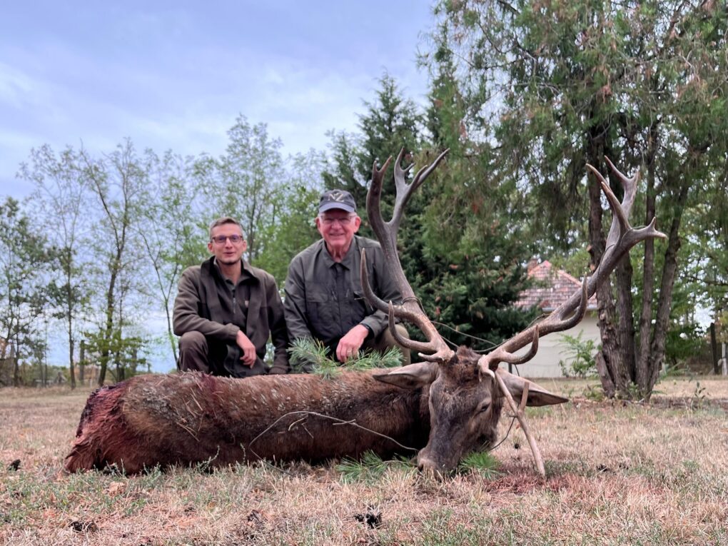 Raymond Bennett — Hungary Red Stag Hunt (OI client photo 2) » Outdoors International Two men crouch beside a large deer with impressive antlers in a yard, posing for a photo outside.