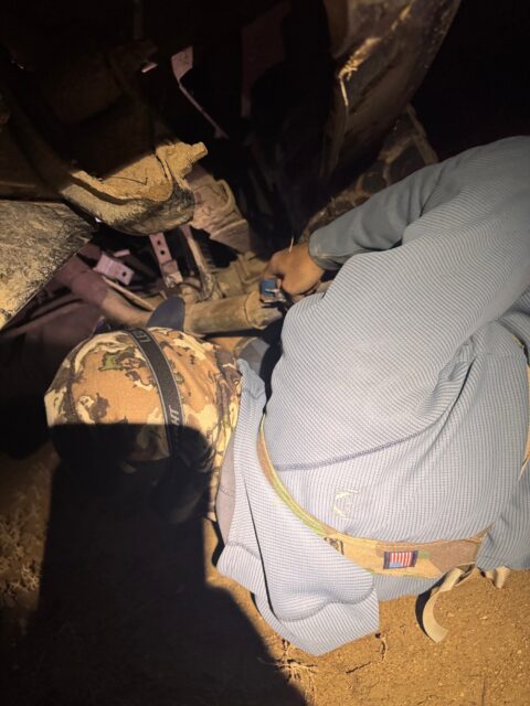 A service member in camouflage gear kneels in a dark, dusty, rubble-filled space, reaching toward a small blue item in the debris.