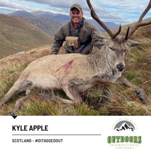 Man in outdoor gear kneels beside a large deer on a grassy mountain hillside, with a small red wound on the deer's side and distant peaks in the background.