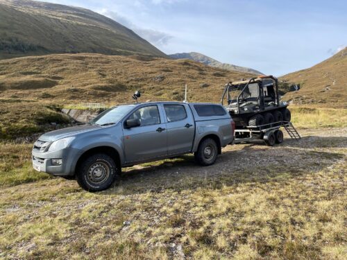 Blue pickup truck towing a multi-track off-road utility vehicle on a trailer in a grassy hillside landscape.