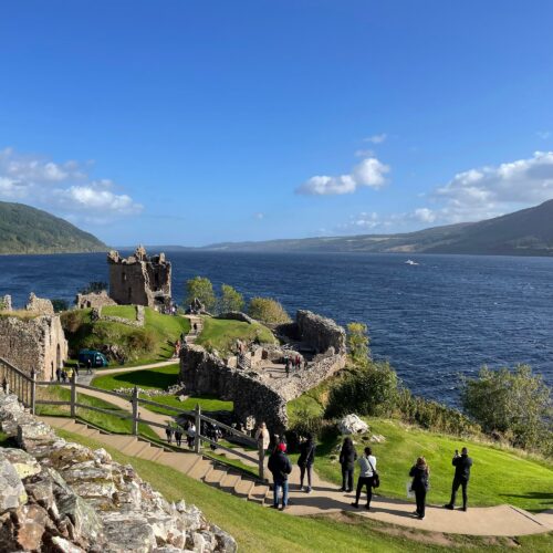 Ruined stone castle by a blue lake, with visitors walking along a path and green lawns in the foreground.