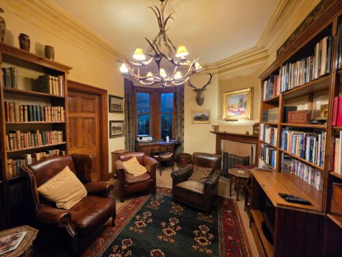 Cozy library room with leather armchairs, tall bookshelves, and an antler chandelier overhead.