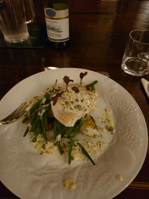 White plate with creamy herb-coated fish and green beans; a wine bottle and glass sit on a dark table in a dim dining room.