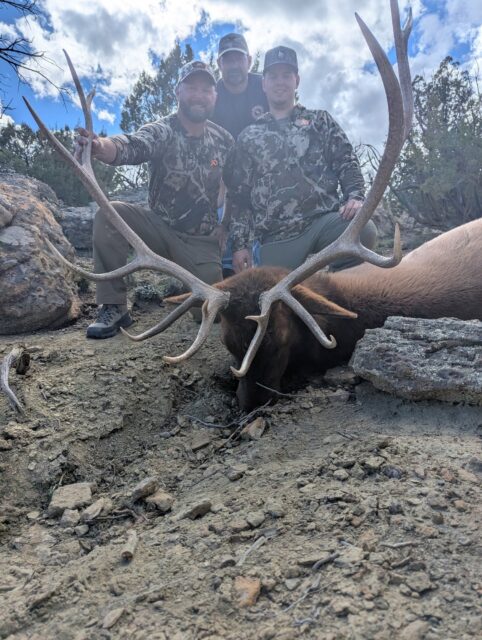 Three hunters in camouflage pose behind a large elk with massive antlers lying on rocky ground in a rugged outdoor setting.