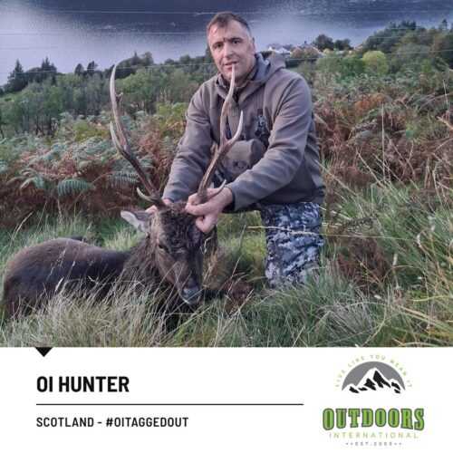 Hunter kneels beside a large deer with broad antlers in a grassy field, Scotland.