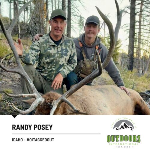 Two hunters posing with a large elk in a forest clearing, showing its broad antlers and the outdoor gear they wear.