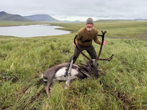Man wearing a beanie and olive shirt poses with a large deer carcass in a grassy field, with mountains and a lake in the distance.