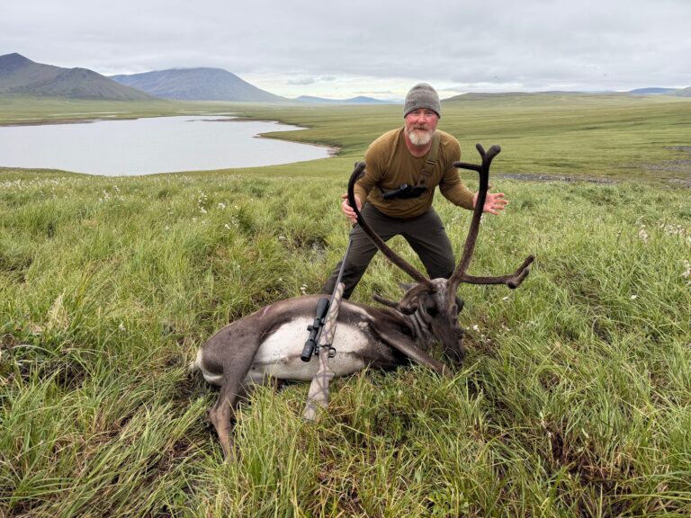 Man wearing a beanie and olive shirt poses with a large deer carcass in a grassy field, with mountains and a lake in the distance.
