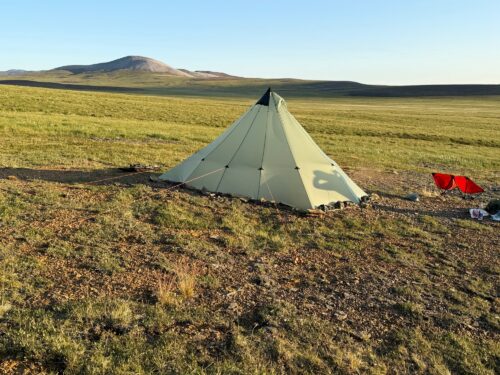 Green pyramid tent pitched on a grassy plain with distant hills and a blue sky, a red folding chair nearby on the right side.